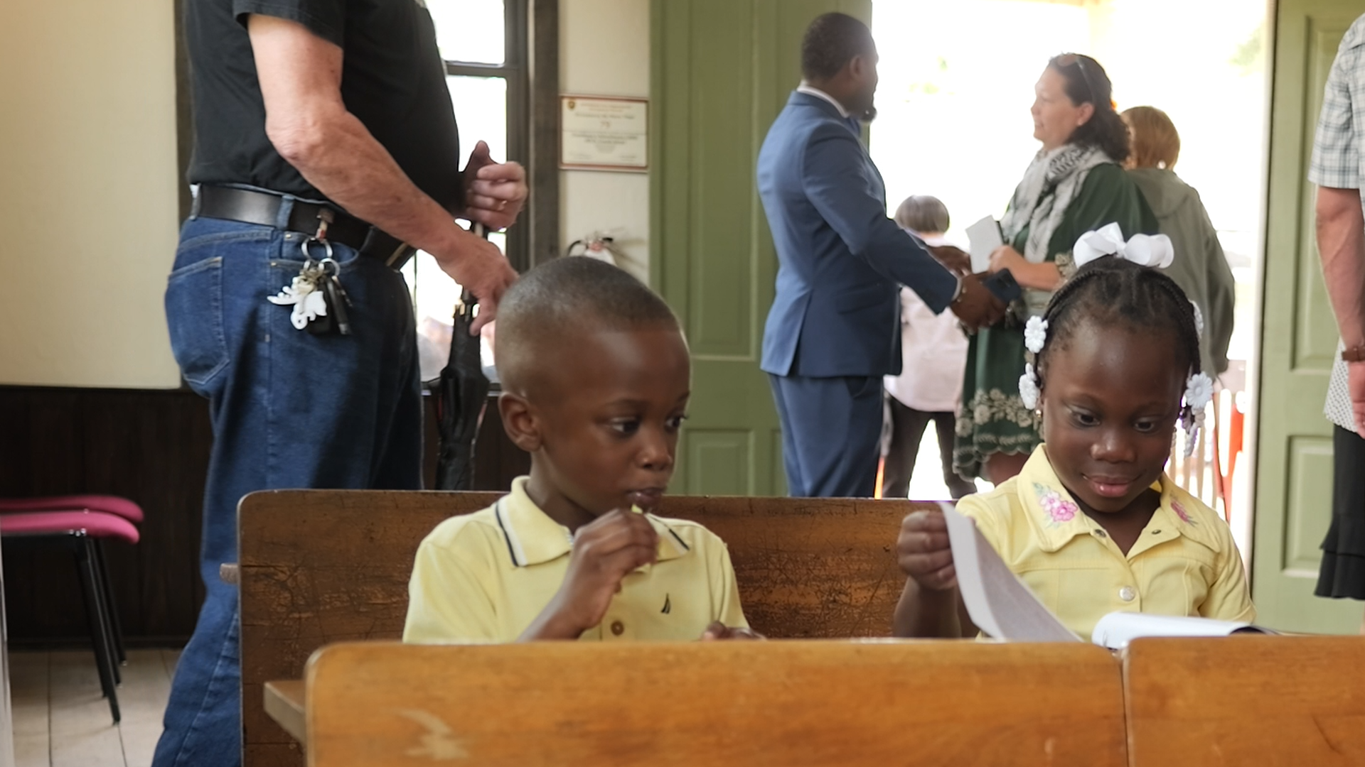 Two children sit at a wooden pew, each holding sheets of paper and looking down at them.